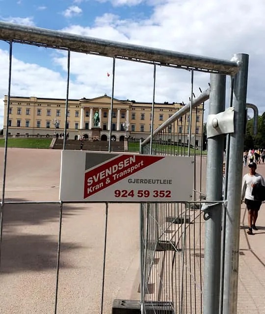 Gjerde med Svendsen Kran & Transport-skilt foran Det kongelige slott i Oslo på solrik dag.